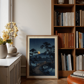 Print leaning on floor of moonlit bridge art print with pine trees and a yellow moon by a bookshelf