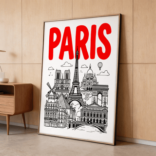 Print leaning on a floor against a light wood wall showing Paris landmarks illustration with red text and a wooden cabinet nearby