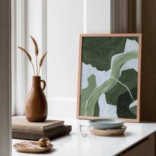 Framed Abstract Green Art Print with abstract green flow and light blue shapes leaning on a white surface near a window vase with dried