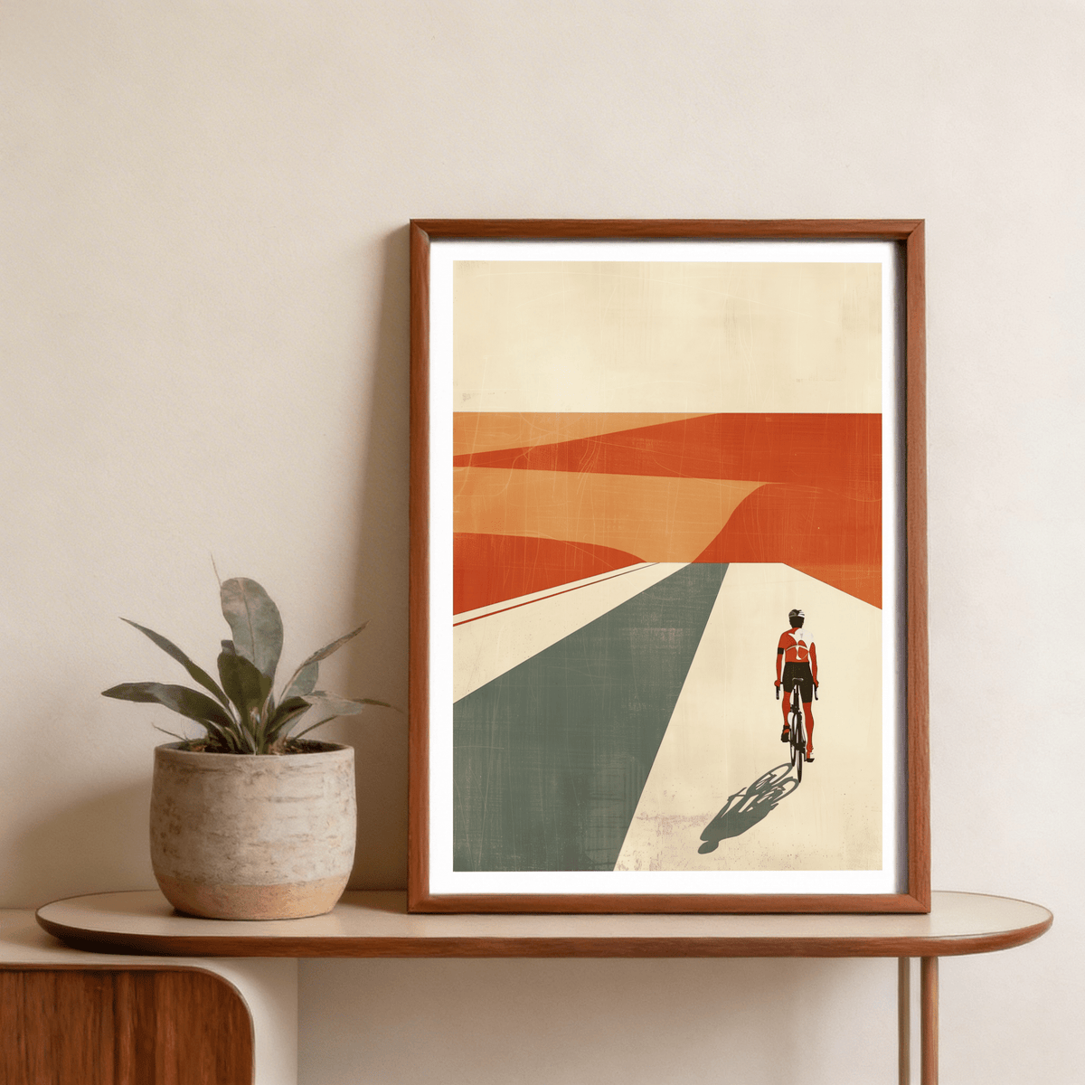Framed Cycling Landscape Print with cyclist in orange and green desert tones beside a plant on a wooden shelf