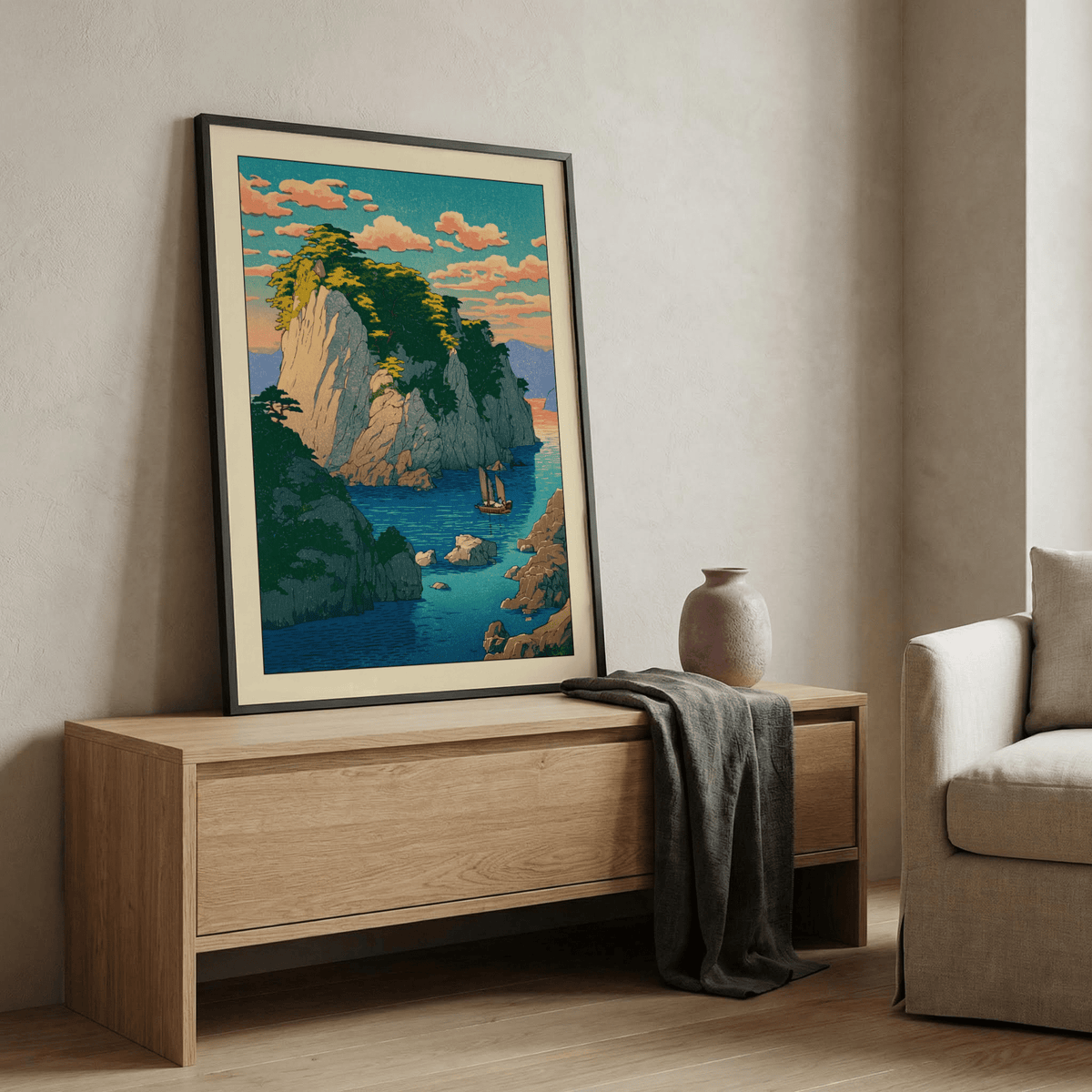 Framed print leaning on a wooden sideboard showing a rocky island with boat in blues and greens with a grey vase nearby
