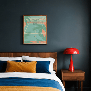 Miami Beach Tennis print on a dark wall above a bed with blue and mustard cushions and a red lamp