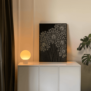 Print leaning on a shelf with dandelions wall art in white and black next to a lit round lamp