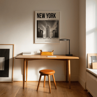 Empire State Building wall art above a wooden desk with an orange pencil holder and brown stool in a softly lit room