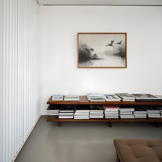 Japanese Cranes Flying Art print above a wooden bench with stacks of books in a minimal room