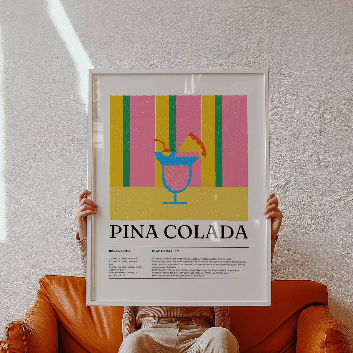 Framed print of piña colada cocktail illustration with colorful stripes held above a brown leather sofa
