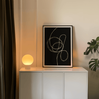 Modern black and white art leaning on a white cabinet with a glowing round lamp nearby