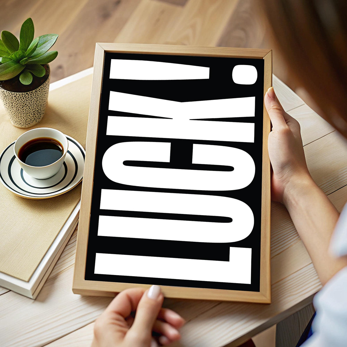Framed print close-up of the typographic word luck in white on black held over a wooden table with a cup of coffee and a plant