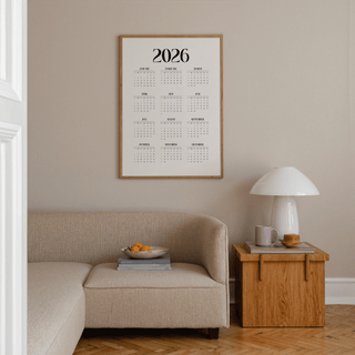 Minimalist wall calendar with black text and orange highlights above a beige sofa and wooden side table with a lamp