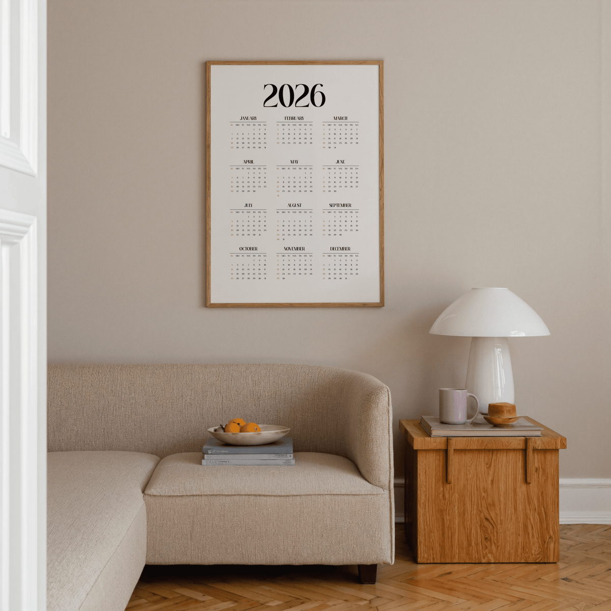 Minimalist wall calendar with black text and orange highlights above a beige sofa and wooden side table with a lamp