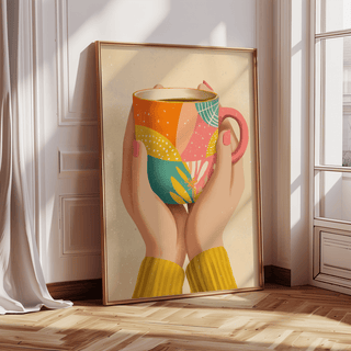 Print leaning on a wooden floor showing hands holding a colourful patterned coffee mug in natural light