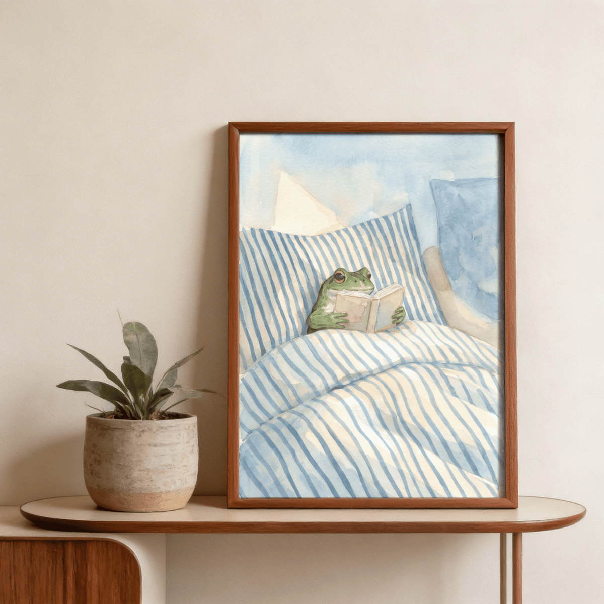 Framed Frog Reading In Bed Print on a wooden table with a plant beside it