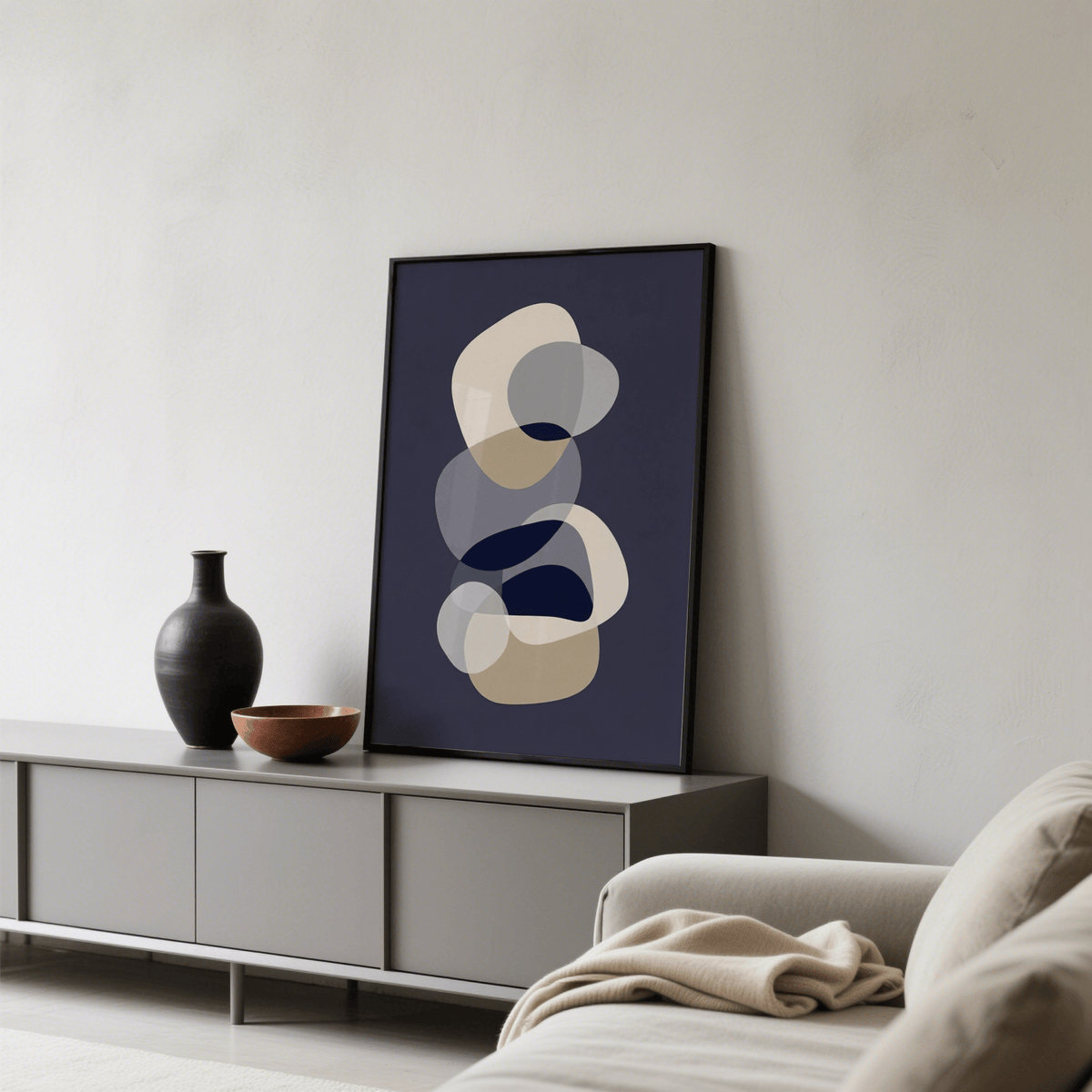 Abstract modern art print with overlapping beige and grey shapes leaning on cabinet beside black vase and bowl