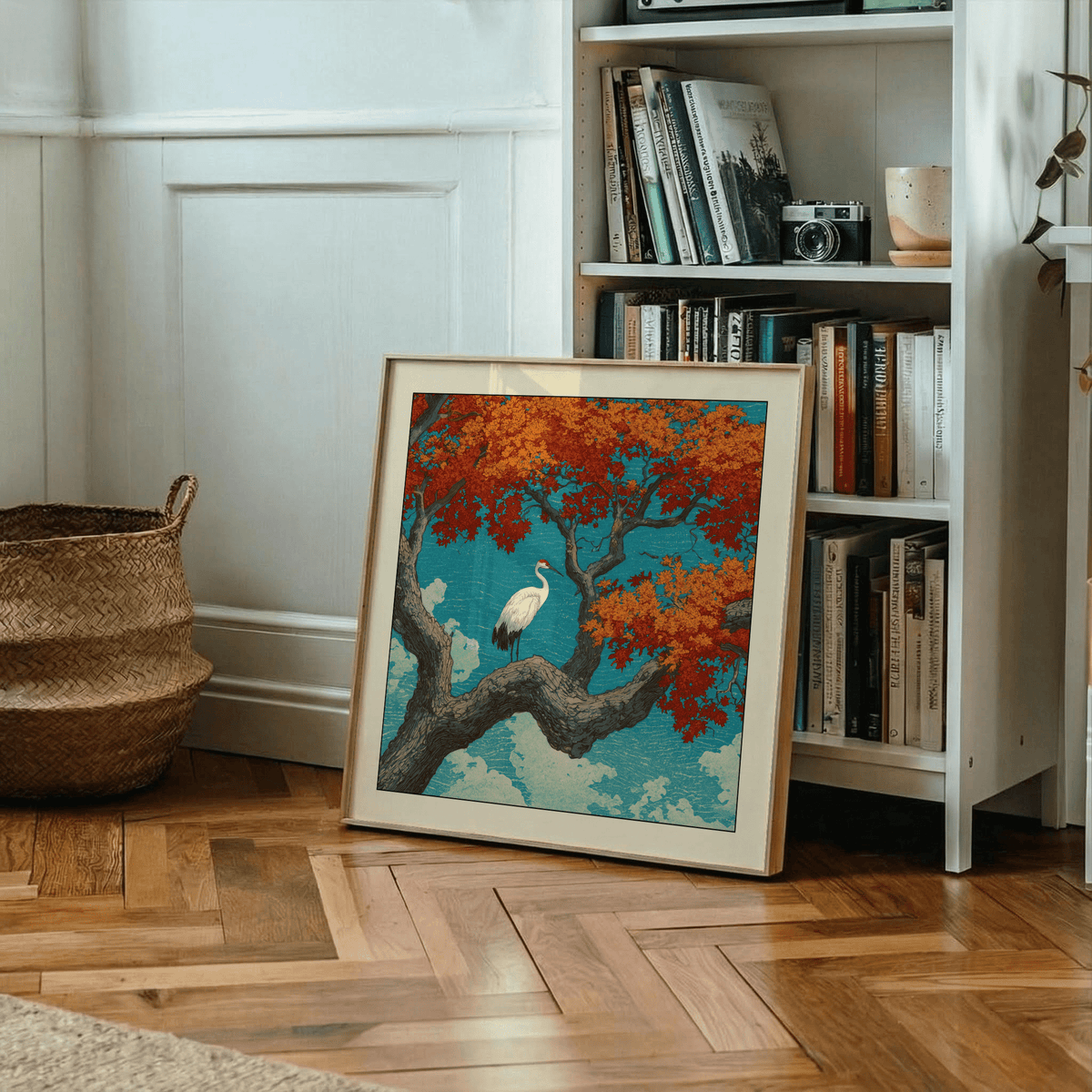 Print leaning on a floor with wooden parquet of a white crane on a tree with red leaves beside a bookcase and woven basket