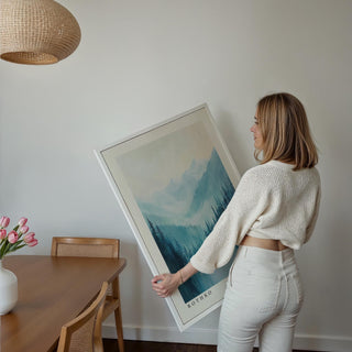 Framed nature print of pine trees and blue mountains held by person near dining table and pink tulips