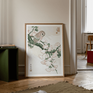 Owl art print leaning on floor with beige ginkgo leaves and green branch beside green table in a living room
