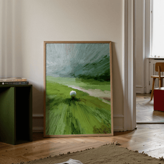 Print leaning on the floor with abstract green golf ball landscape and light wood frame beside a stack of books