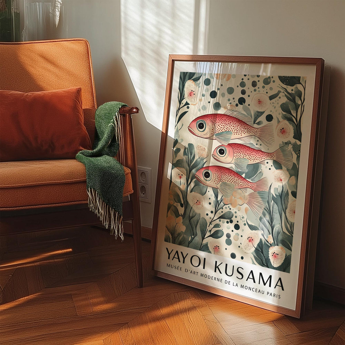 Fish artwork print leaning on a wooden floor near an orange chair and green throw showing red spotted fish in botanical style