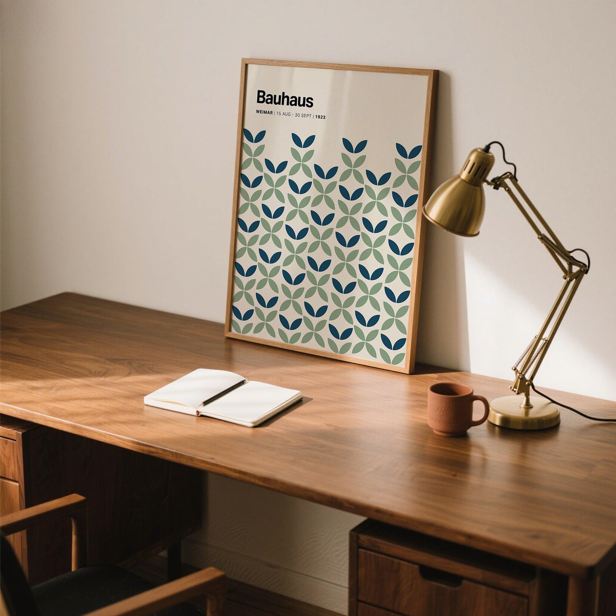 Print leaning on a shelf with geometric blue and green leaf pattern beside a brass lamp on a wooden desk