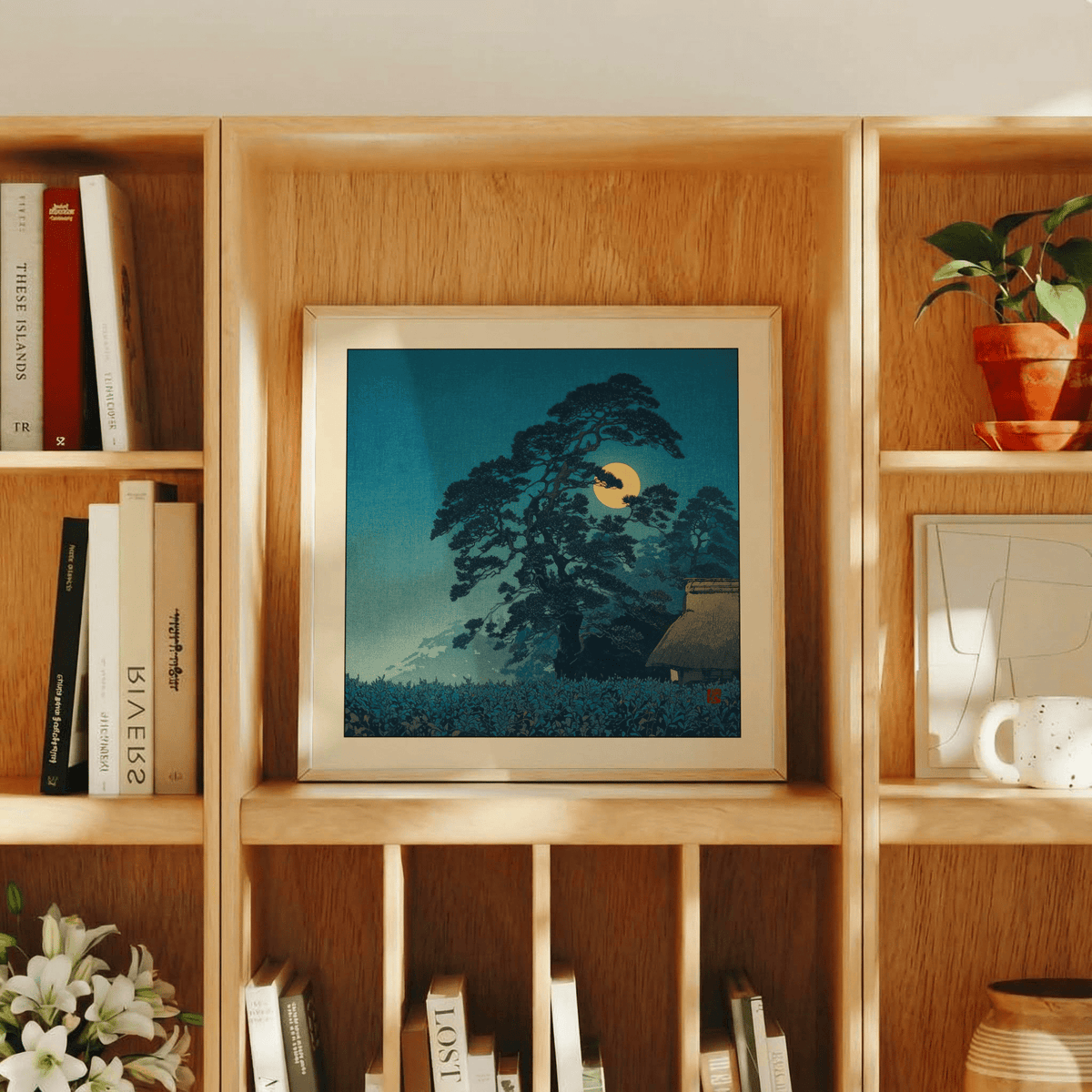 Print of Hasui Kawase Moon Print with a pine tree and full moon in blue and yellow tones on a wooden shelf with books and plant