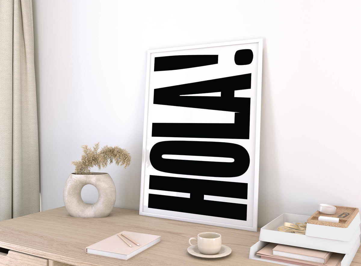 Print leaning on a desk of typographic posters with bold black HOLA text and white frame next to a vase