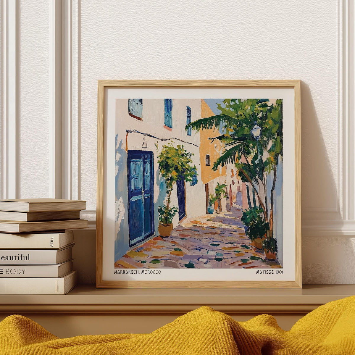 Framed Morocco wall art of a colourful Mediterranean street scene with a blue door leaning on a shelf by books
