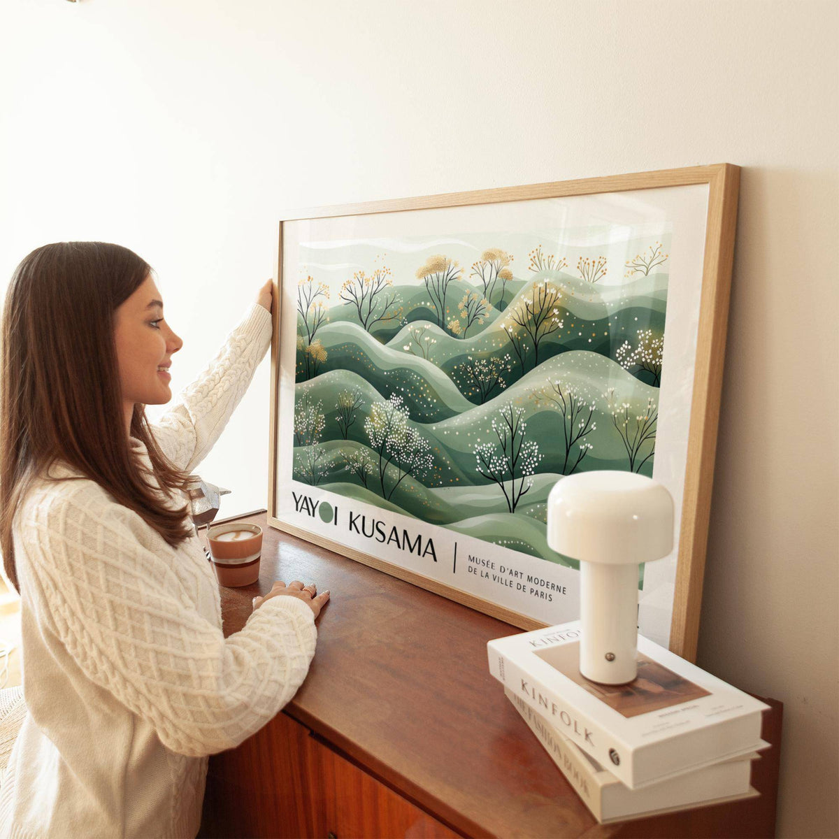 Landscape art prints leaning on a wooden sideboard with minimalist green hills and blossoming trees next to a white lamp