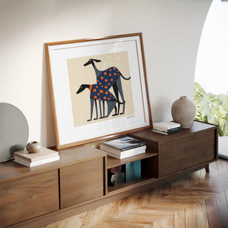 Framed print of two greyhounds with orange polka dots leaning on a wooden sideboard with books and pottery