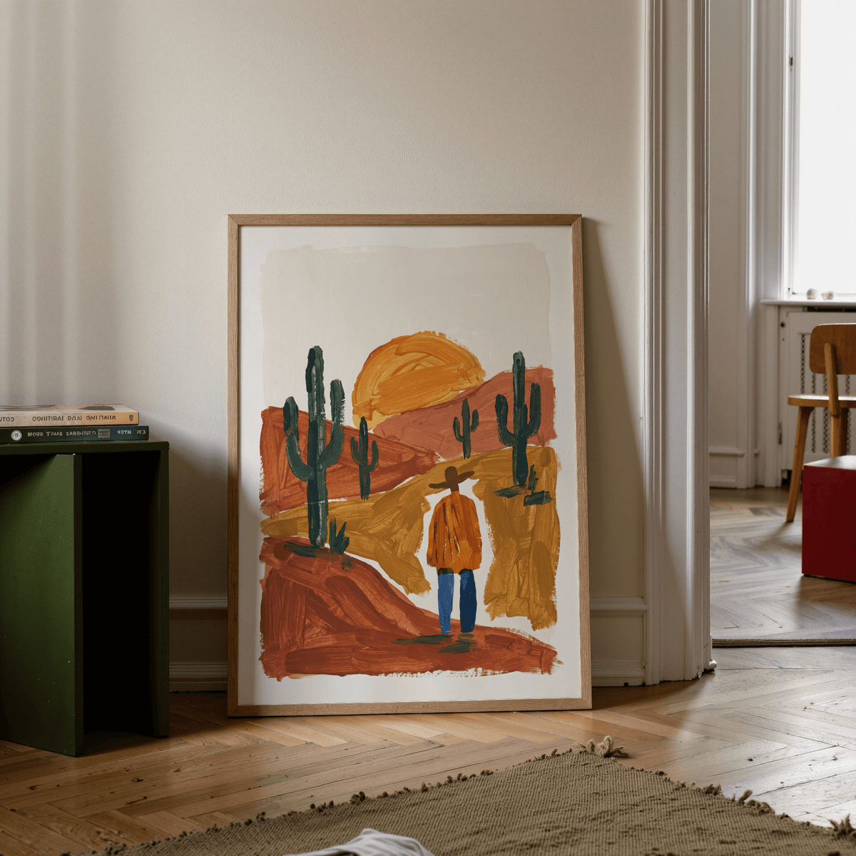 Print leaning on floor depicting desert landscape art print with abstract man and cacti near green shelf