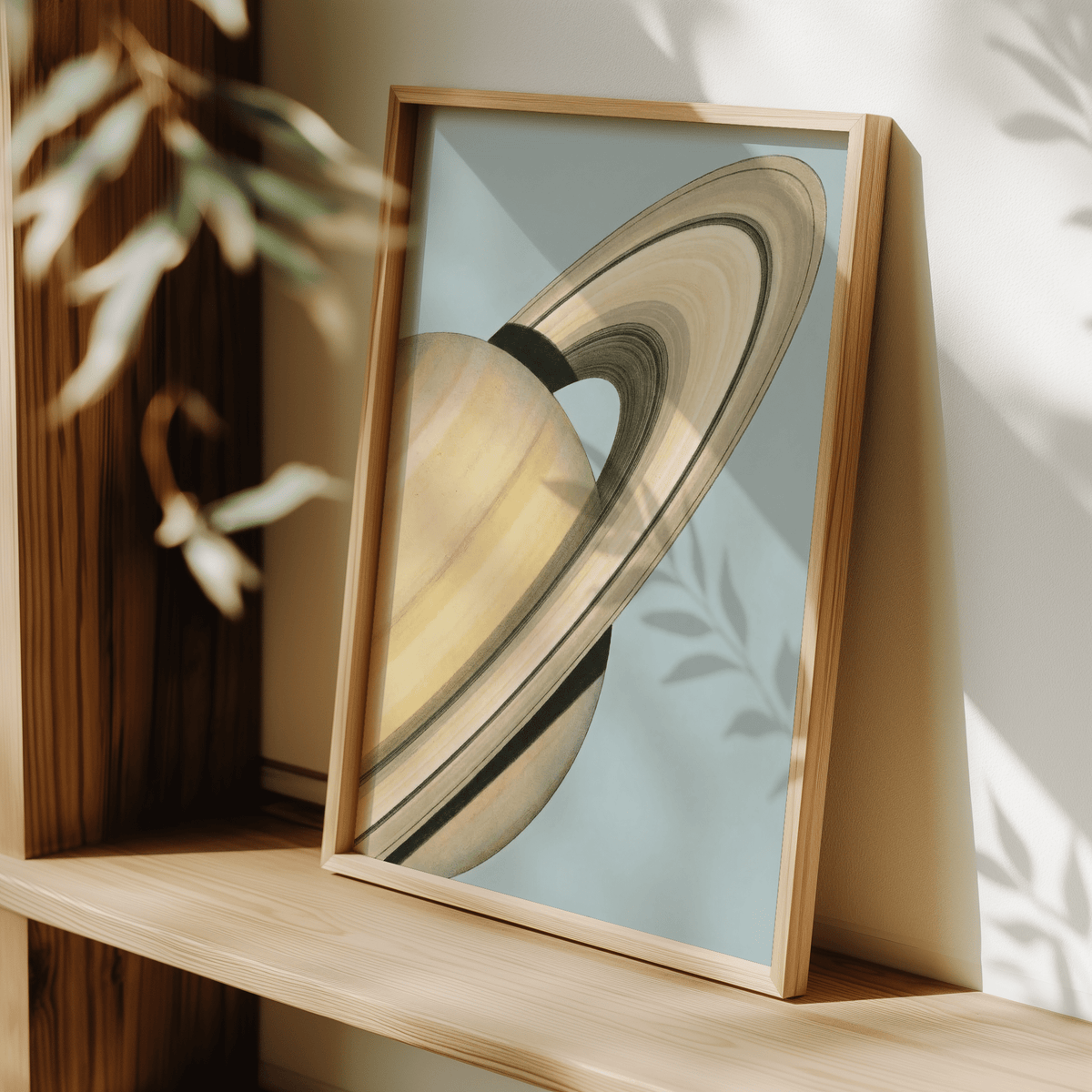 Framed Saturn wall art with beige and blue minimalist illustration leaning on a wooden shelf with plant shadows
