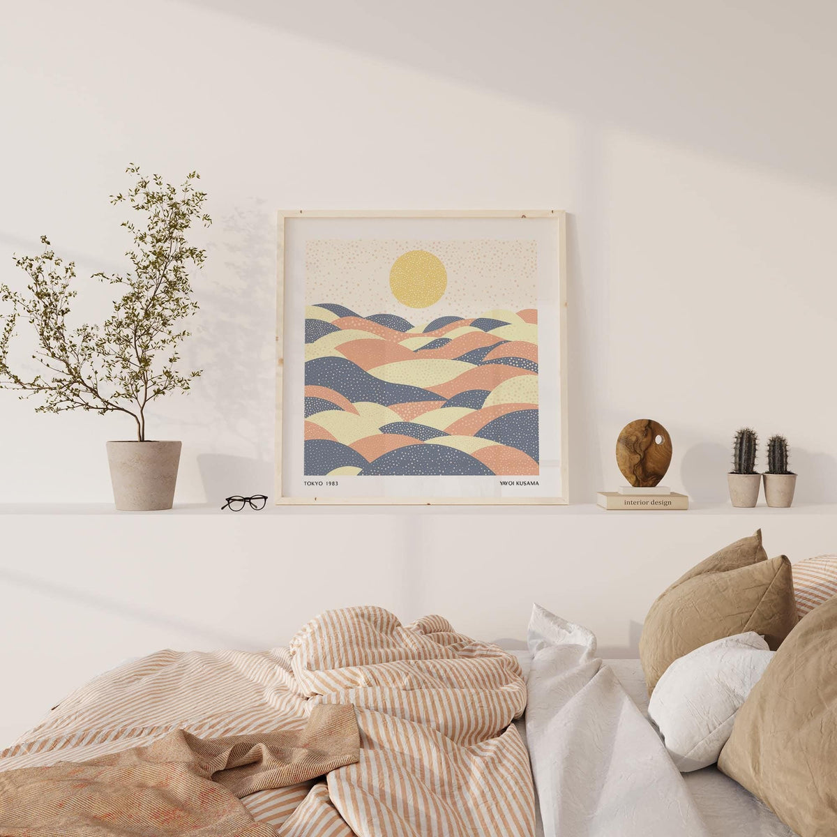 Print leaning on a shelf of abstract hills with a yellow sun in pastel colours above a bed with cushions