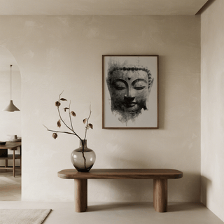 Framed print of Buddha face in black and grey watercolour above a wooden table with a large glass vase in a neutral toned room