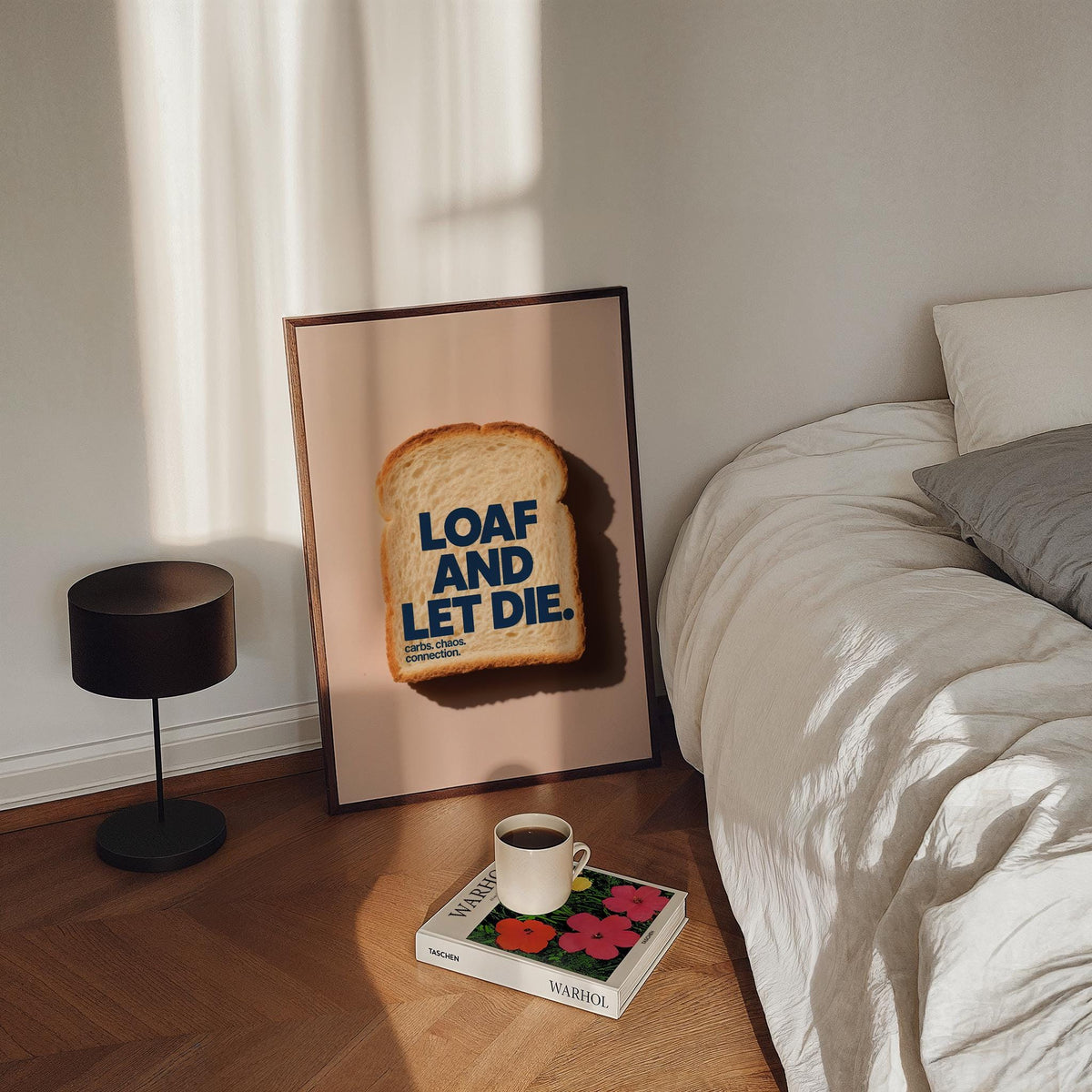 Quirky Kitchen Art print leaning on floor beside a bed with a cup on a book and black lamp nearby
