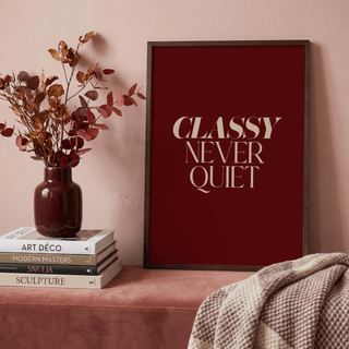 Classy wall art with beige text on deep red background leaning on a mauve surface next to a vase with dried flowers
