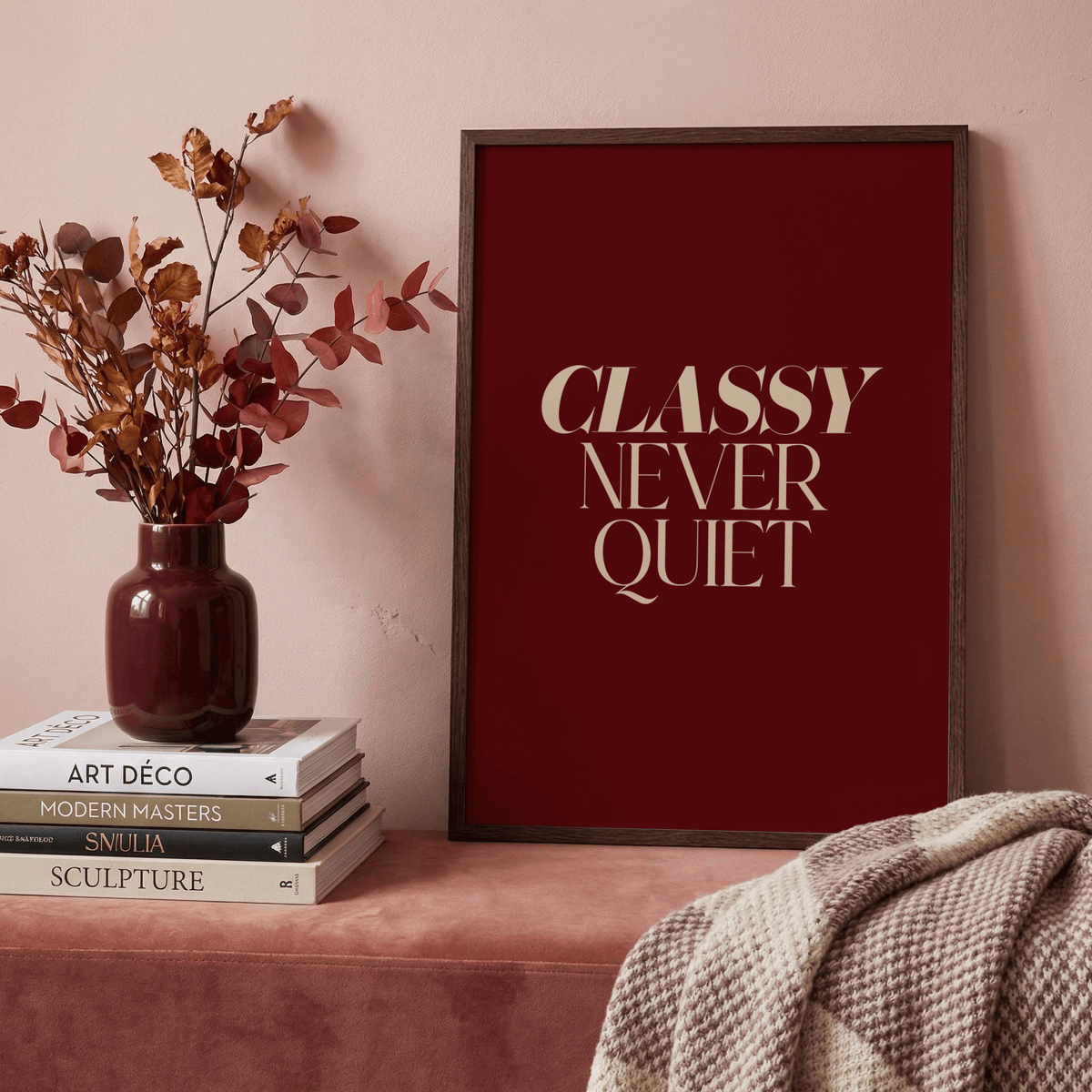 Classy wall art with beige text on deep red background leaning on a mauve surface next to a vase with dried flowers