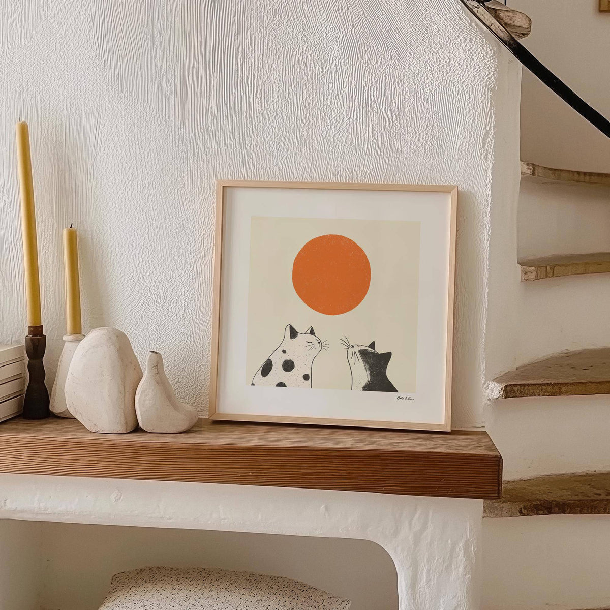 Print leaning on a shelf featuring two black and white cats with an orange sun and beige background next to candles