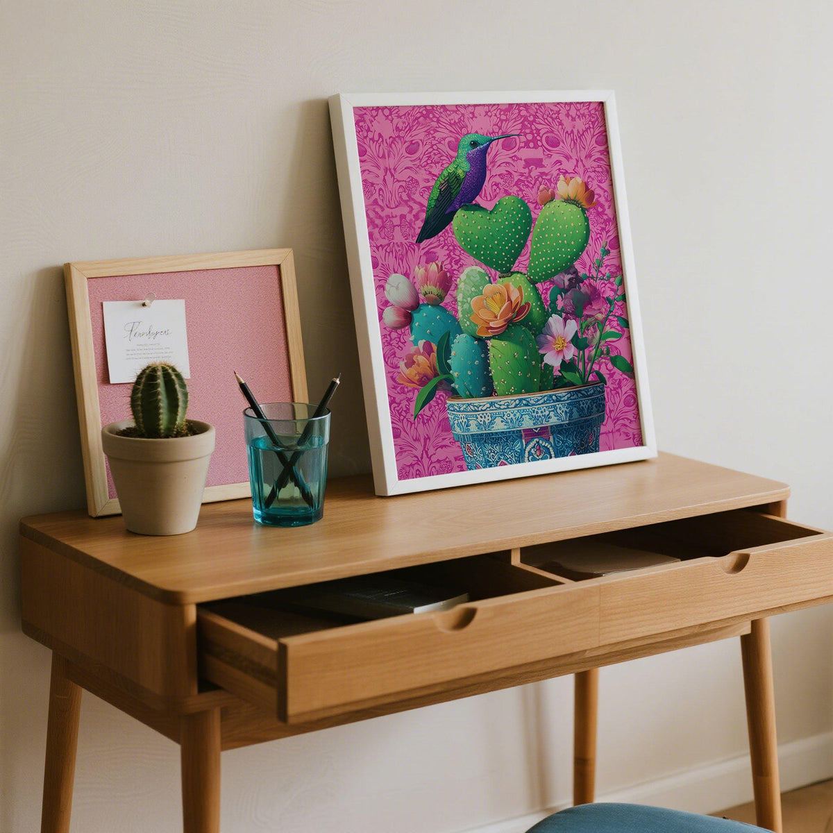 Print leaning on a shelf of a heart shaped cactus with a hummingbird on a pink background beside a small potted cactus
