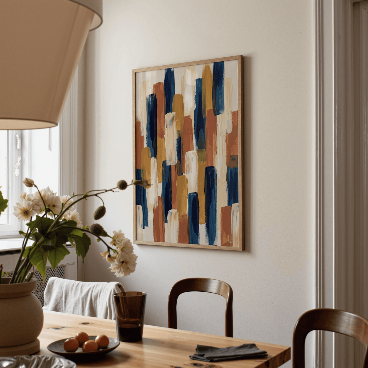 Print on a wall of abstract color blocks in blue, brown and cream beside a dining table with a lamp and flowers