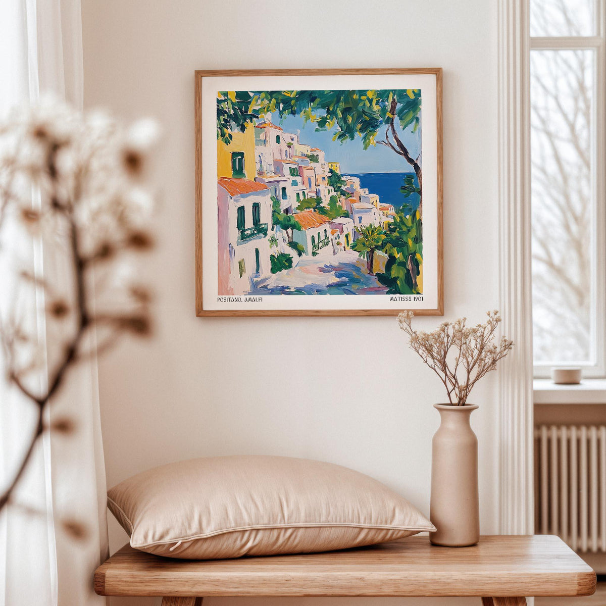 Print on a wall in a light room of Positano art showing colourful buildings by the sea with green and blue tones with a vase nearby