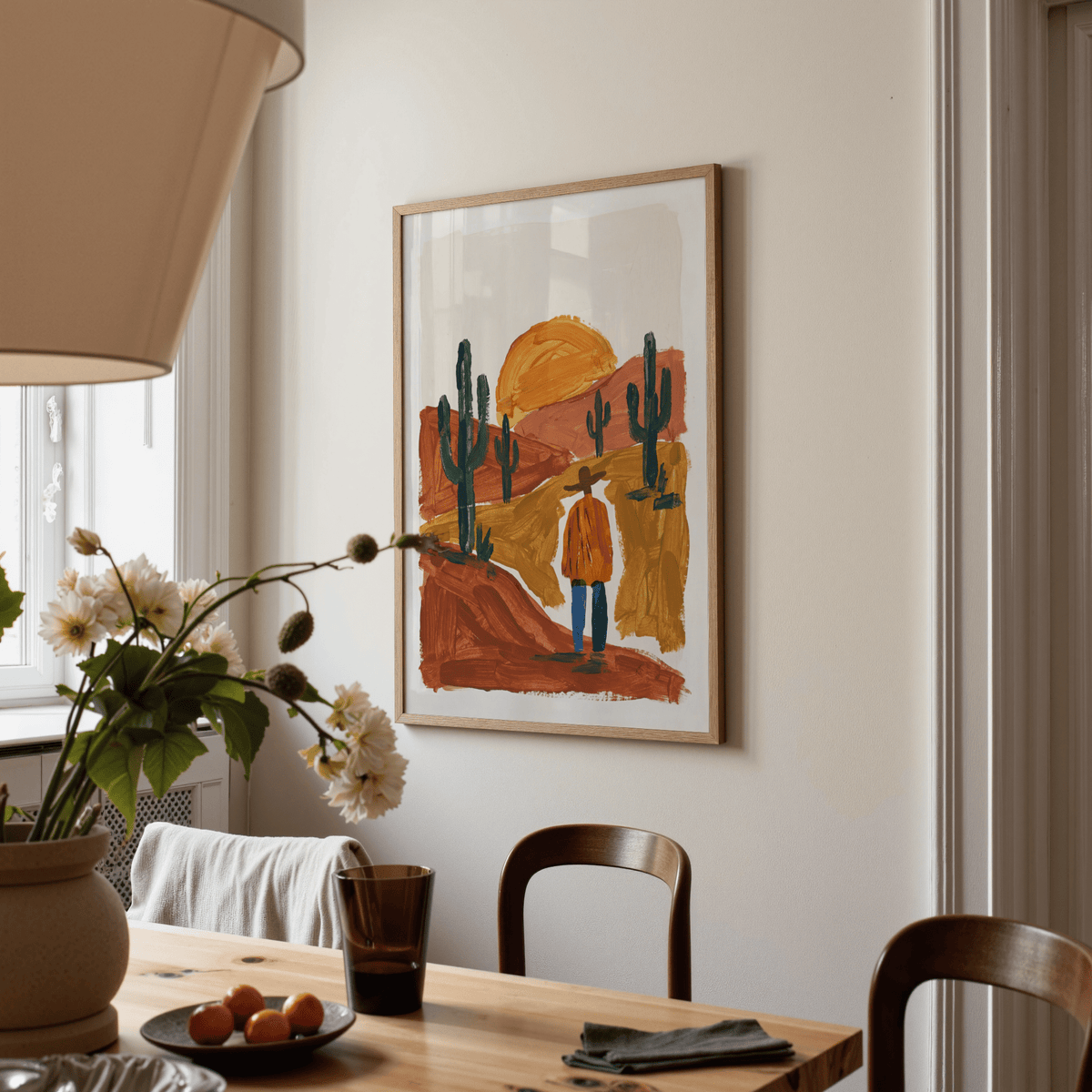 Print on a wall in a dining room showing a man in a desert with orange sun and cacti with flower vase on table