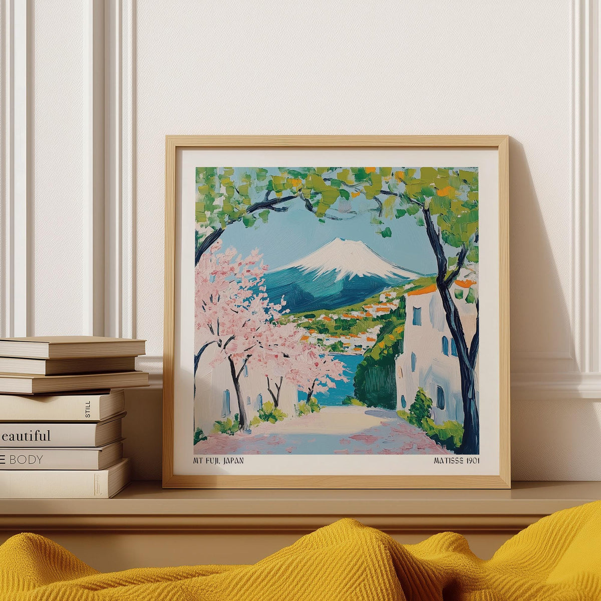 Framed Japan poster of Mount Fuji with pink cherry blossoms and green leaves leaning on a shelf next to books