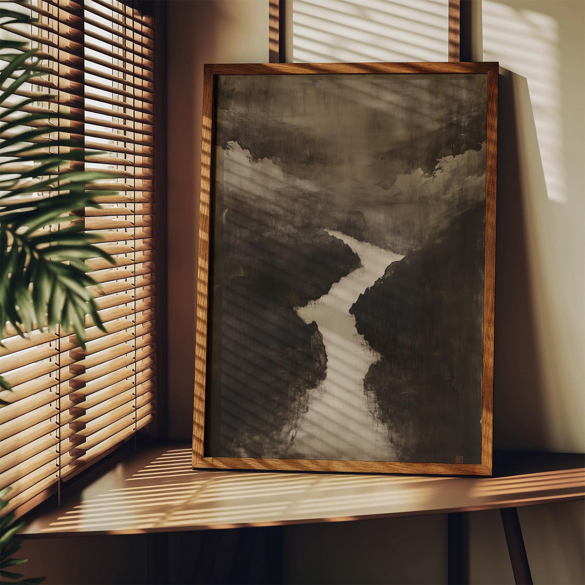 Framed Sumi e Art of a river winding through a minimalist landscape in soft greys leaning on a wooden shelf with plant leaves nearby