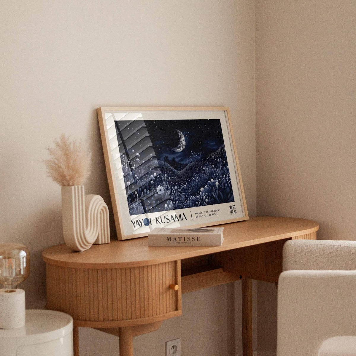 Print leaning on a desk of a crescent moon over floral hills with dark blue and white tones beside a beige vase