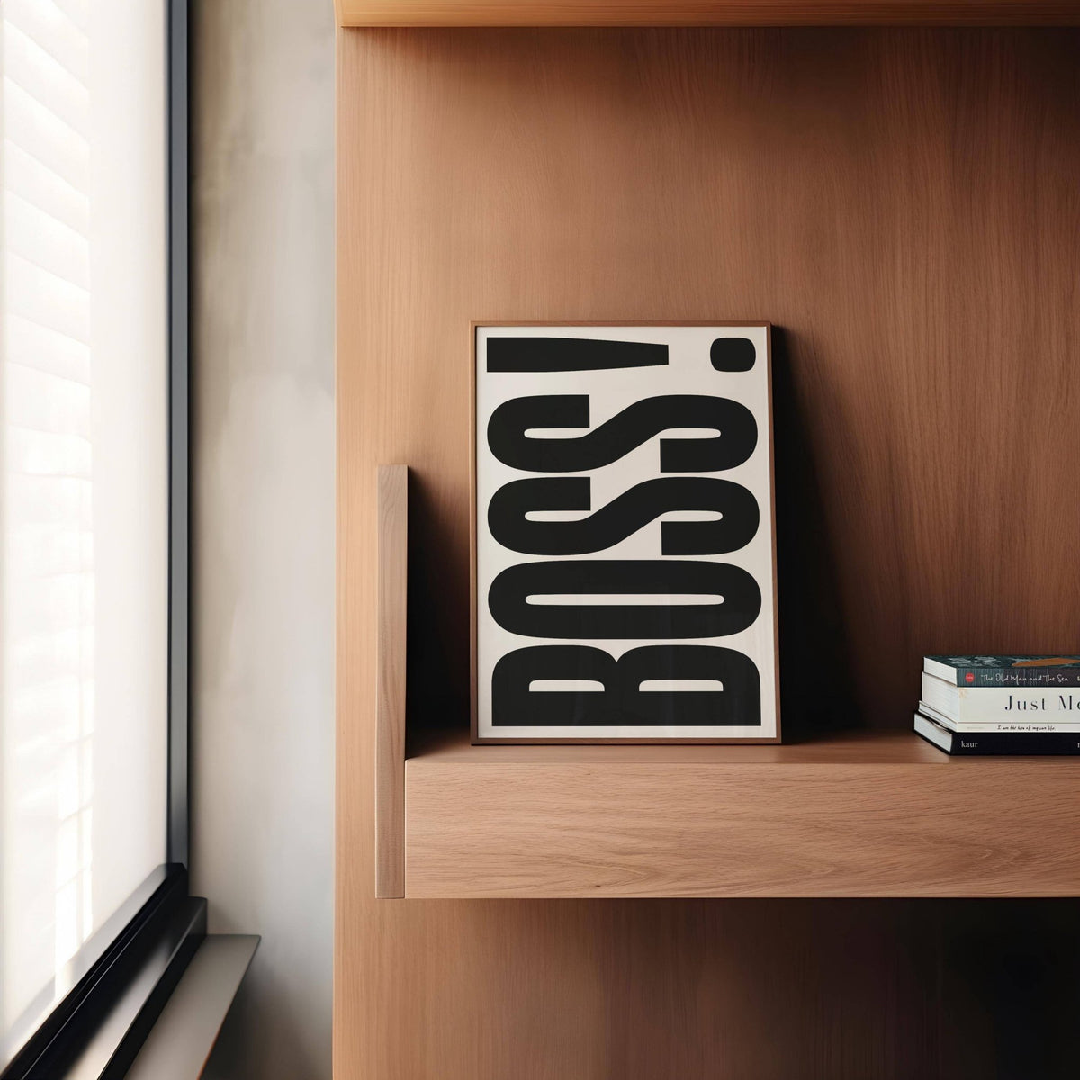 Office wall art with boss typography in black and white leaning on a wooden shelf next to books and a window