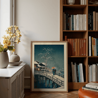 Print leaning on floor of snowy bridge with two people in traditional dress beside bookshelves and plants