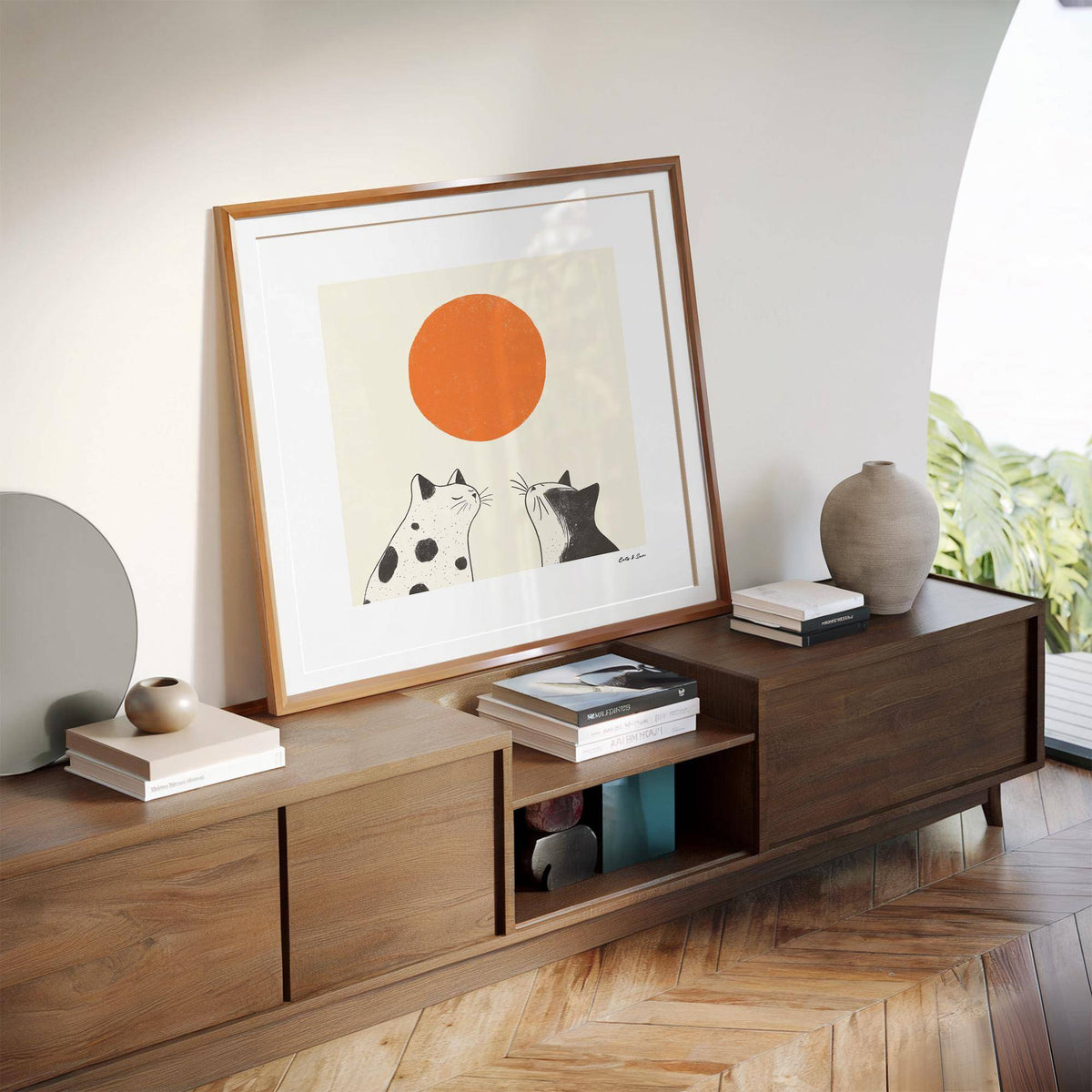 Print leaning on a shelf showing minimalist illustration of two black and white cats with an orange sun and neutral background