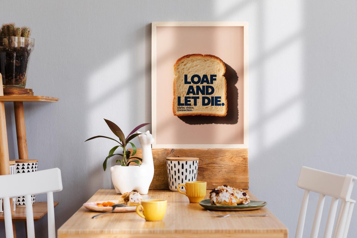Quirky Kitchen Art print of a slice of bread with blue text on a pink background above a wooden dining table with mugs and a plant