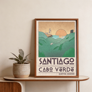 Framed Santiago Cabo Verde art print illustrating a sunset over coastal hills with green and orange tones placed on a wooden table next to