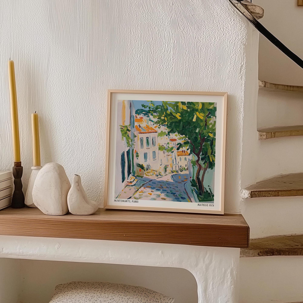 French artwork prints leaning on a wood shelf beside candles and a fruit sculpture in a white room with stairs