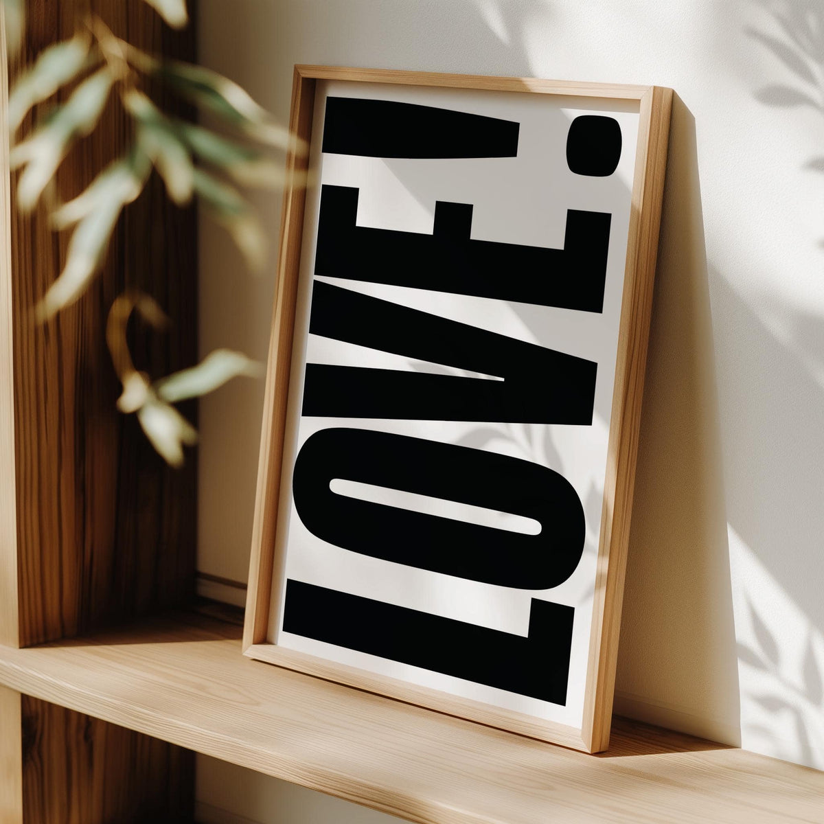 Framed LOVE typography poster with black text on white leaning on a wooden shelf with leaves nearby